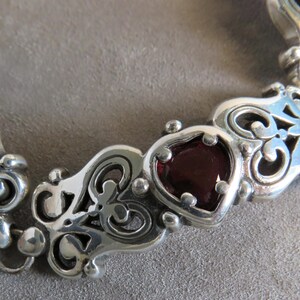 Pu&ograve; includere: Un bracciale a maglie in argento caratterizzato da motivi ornamentali in metallo traforato a volute, che si alternano a gemme rosso scuro levigate a forma di cuore. Il bracciale &egrave; composto da pi&ugrave; segmenti collegati, ognuno con dettagli metallici simmetrici.
