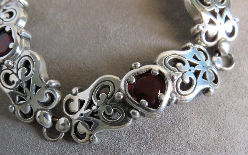 Pu&ograve; includere: Un bracciale a maglie in argento caratterizzato da motivi ornamentali in metallo traforato a volute, che si alternano a gemme rosso scuro levigate a forma di cuore. Il bracciale &egrave; composto da pi&ugrave; segmenti collegati, ognuno con dettagli metallici simmetrici.