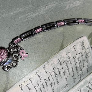 Peut inclure: Bracelet argent&eacute; avec des d&eacute;tails et des breloques roses. Le bracelet comporte un charm en forme de c&oelig;ur recouvert de cristaux roses et un charm ruban rose. Le bracelet est pr&egrave;s d'une note manuscrite avec du texte visible.