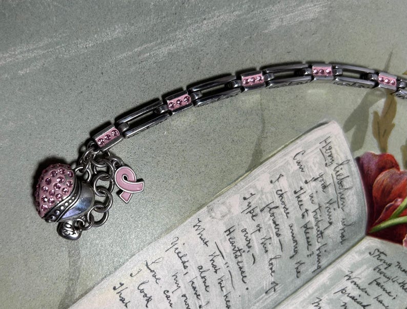 Peut inclure: Bracelet argent&eacute; avec des d&eacute;tails et des breloques roses. Le bracelet comporte un charm en forme de c&oelig;ur recouvert de cristaux roses et un charm ruban rose. Le bracelet est pr&egrave;s d'une note manuscrite avec du texte visible.