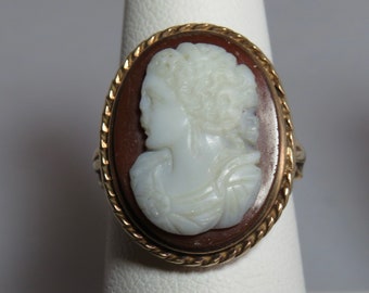 May include: A gold ring with a cameo setting. The cameo is a white cameo of a woman's profile with a brown background.