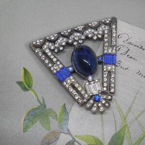 May include: A triangular brooch featuring a large oval dark blue cabochon stone at the center, accented with smaller round blue stones, rectangular blue baguettes, clear rhinestones, and a clear baguette stone set in a silver-toned metal frame.