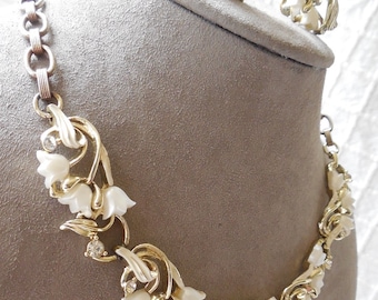 May include: A gold-tone metal necklace and matching clip-on earrings set featuring white floral lily-of-the-valley motifs accented with small clear rhinestones. The necklace has a link chain and the earrings mirror the floral design of the necklace.