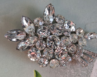 May include: A silver brooch featuring a cluster of clear rhinestones in various shapes and sizes. The brooch has a decorative bow detail.