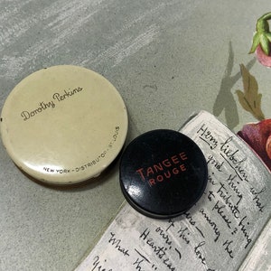 May include: A round, cream-colored Dorothy Perkins compact with black text reading New York - Distributor - St. Louis and a smaller, round, black Tangee Rouge compact with red text, both resting on a surface with floral illustrations and script.