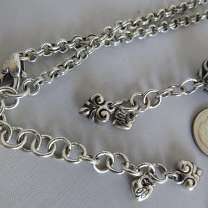 May include: A silver chain necklace with multiple charms, including a heart, a swirl design, and a larger round charm with intricate details. The necklace has a lobster claw clasp.