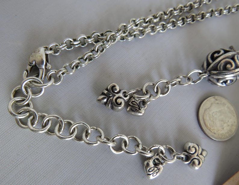 May include: A silver chain necklace with multiple charms, including a heart, a swirl design, and a larger round charm with intricate details. The necklace has a lobster claw clasp.