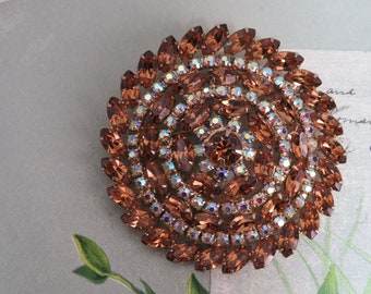 May include: A large, round brooch made of brown and iridescent rhinestones. The rhinestones are arranged in a spiral pattern, creating a swirling effect.