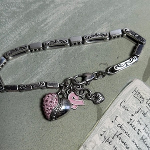 Peut inclure: Bracelet argent&eacute; compos&eacute; de maillons rectangulaires et de volutes d&eacute;coratives. Il comporte des breloques, dont un c&oelig;ur orn&eacute; de cristaux roses, un ruban rose et un petit c&oelig;ur. Un texte manuscrit est visible en arri&egrave;re-plan.