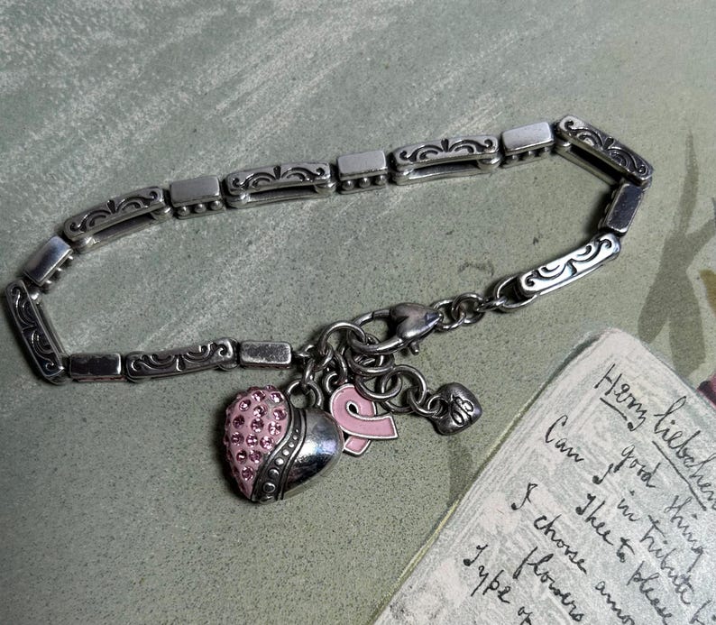 Peut inclure: Bracelet argent&eacute; compos&eacute; de maillons rectangulaires et de volutes d&eacute;coratives. Il comporte des breloques, dont un c&oelig;ur orn&eacute; de cristaux roses, un ruban rose et un petit c&oelig;ur. Un texte manuscrit est visible en arri&egrave;re-plan.