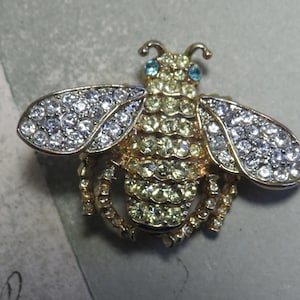 May include: A gold-toned bee brooch encrusted with clear and yellow rhinestones. The wings are detailed with clear rhinestones, and the eyes are small blue stones. This jewelry item is a decorative accessory.