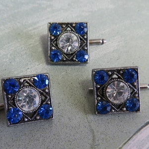 May include: Three silver cufflinks with a square design. Each cufflink has a large clear rhinestone in the center surrounded by four smaller blue rhinestones.
