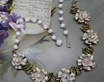 May include: A necklace featuring a series of white spherical beads connected to a chain of five white enamelled flower charms with green enamelled leaves and iridescent central crystals. The necklace includes a gold-toned metal hook clasp closure.