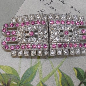May include: A rectangular two-piece dress clip featuring a silver-tone metal frame encrusted with clear and pink rhinestones. The design includes geometric patterns and rows of sparkling stones arranged in a symmetrical, elongated, and notched shape.