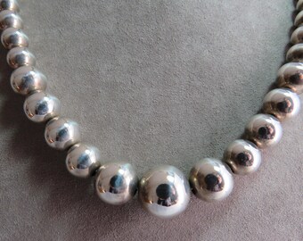 May include: A necklace composed of polished silver-toned spherical beads that gradually increase in size toward the center. The beads have a reflective, metallic finish and are strung together in a continuous, curved arrangement.