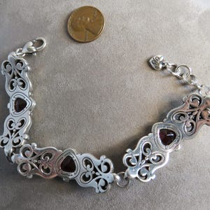 Pu&ograve; includere: Un bracciale a maglie color argento con decorazioni a volute e tre pietre rosso scuro a forma di cuore. Il bracciale include una chiusura a moschettone e un piccolo ciondolo a forma di cuore. Una moneta &egrave; inclusa per dare un'idea della scala.