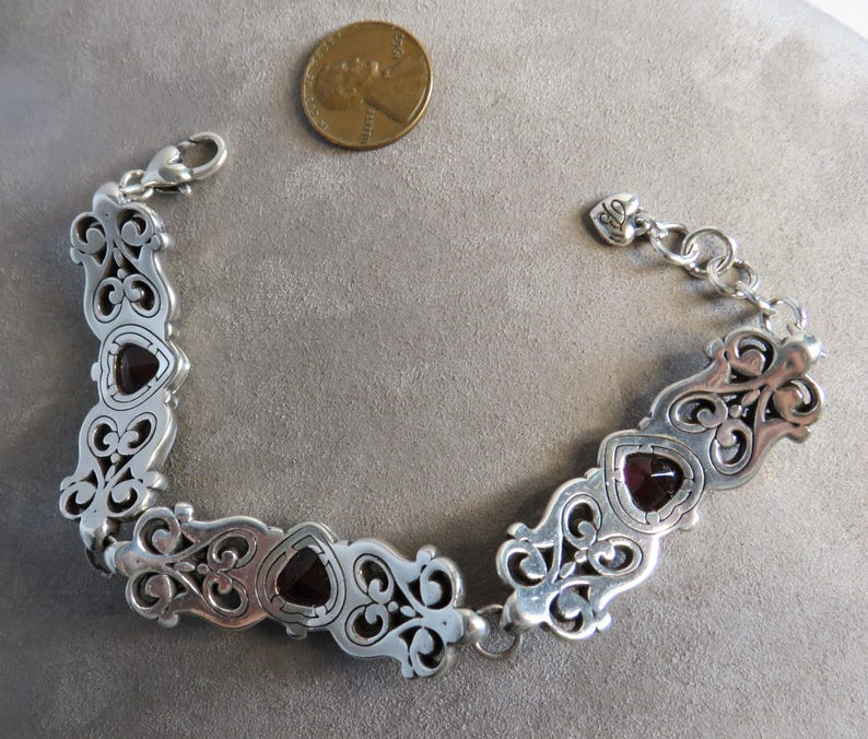 Pu&ograve; includere: Un bracciale a maglie color argento con decorazioni a volute e tre pietre rosso scuro a forma di cuore. Il bracciale include una chiusura a moschettone e un piccolo ciondolo a forma di cuore. Una moneta &egrave; inclusa per dare un'idea della scala.