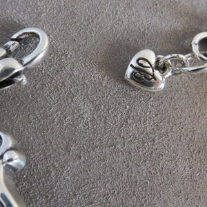 Pu&ograve; includere: Un bracciale in metallo color argento con chiusura a moschettone e un piccolo ciondolo a forma di cuore inciso con le lettere JB. Il bracciale include una catena a maglie circolari e un componente decorativo in metallo in stile filigrana.