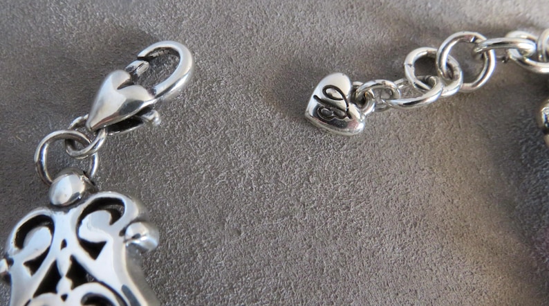 Pu&ograve; includere: Un bracciale in metallo color argento con chiusura a moschettone e un piccolo ciondolo a forma di cuore inciso con le lettere JB. Il bracciale include una catena a maglie circolari e un componente decorativo in metallo in stile filigrana.