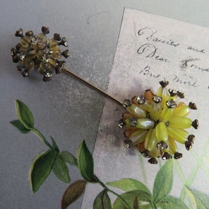 May include: A vintage hat pin with two decorative floral clusters. The clusters feature yellow and clear beads with rhinestone accents, connected by a long, gold-toned pin. The pin is set against a backdrop of a vintage letter.