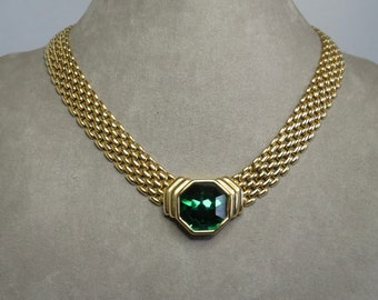 May include: A gold-tone necklace featuring a wide, woven-link chain design. The centerpiece is a large, faceted, octagonal dark green stone set in a polished gold-tone frame that connects to the chain on both sides.