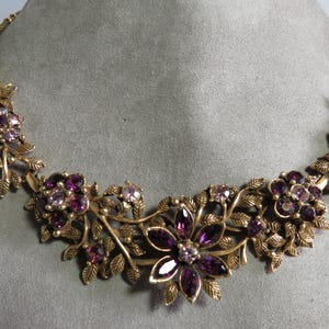 May include: A gold-toned necklace featuring a floral design with purple and lavender-colored gemstones. The necklace has a botanical theme, with leaves and flower clusters. The gemstones are set in a detailed, ornate setting.