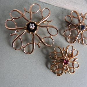 May include: Three gold-toned brooches with intricate designs. The largest brooch features a black center with a clear stone, while the other two brooches have red and blue stones in the center.