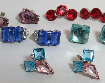 May include: A collection of five pairs of earrings featuring faceted glass stones in various shapes and colours, including light purple, red, blue, and light blue, set in metal frames with screw-back or clip-on mechanisms.