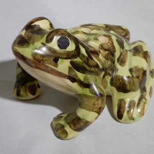 May include: A ceramic frog figurine featuring a glossy finish with a light cream base color and irregular dark brown and olive green painted spots. The frog has a smooth, rounded body, prominent eyes with black pupils, and four distinct legs.
