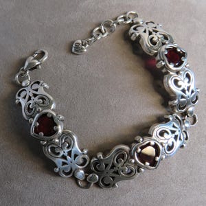 Pu&ograve; includere: Un bracciale a maglie in argento con sezioni alternate in filigrana metallica decorata e pietre rosso scuro a forma di cuore. Il bracciale include una chiusura a moschettone e una catenella di estensione con un piccolo ciondolo a cuore.