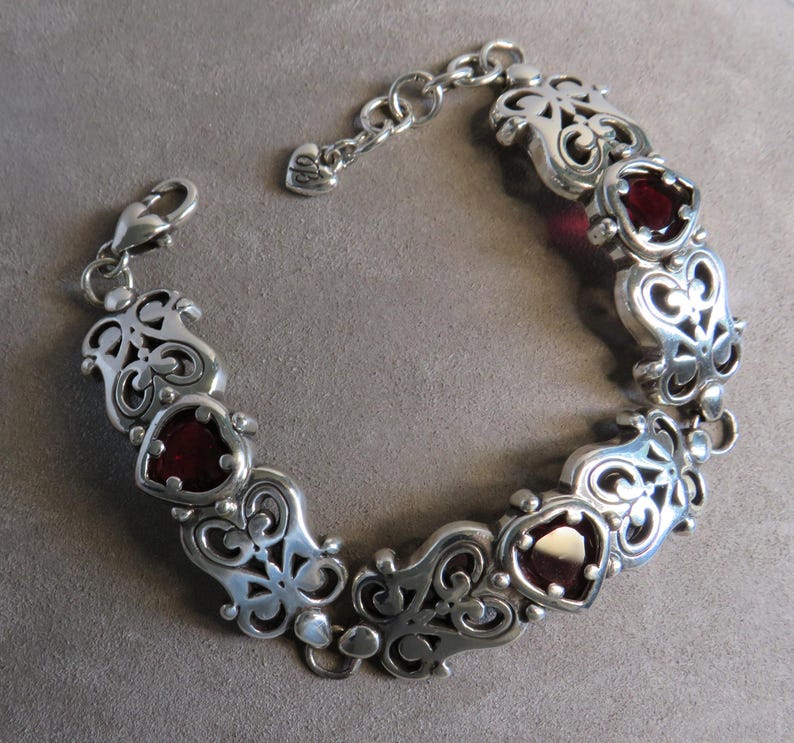 Pu&ograve; includere: Un bracciale a maglie in argento con sezioni alternate in filigrana metallica decorata e pietre rosso scuro a forma di cuore. Il bracciale include una chiusura a moschettone e una catenella di estensione con un piccolo ciondolo a cuore.