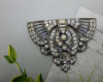 May include: A silver-toned metal brooch featuring a fan-shaped design adorned with numerous clear, round, and rectangular rhinestones. The piece has a central oval frame filled with smaller stones and a symmetrical, intricate metalwork pattern.