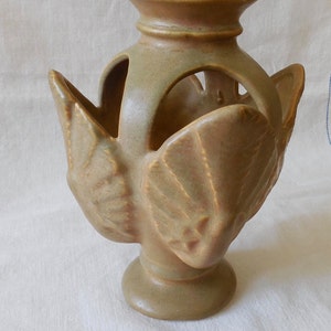 May include: A tan ceramic vase featuring a central cylindrical neck and a flared rim. The body is adorned with four symmetrical, leaf-shaped protrusions that extend outward from the base, creating open arched spaces between the neck and the leaves.