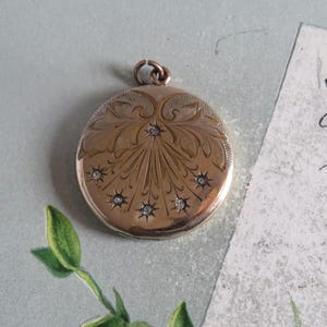 May include: A gold-colored, round locket with an engraved floral design and small, sparkling gemstones. The locket has a loop at the top for a chain. The design features a central burst pattern with radiating lines and decorative flourishes.