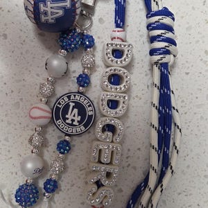 Op de afbeelding: Een Los Angeles Dodgers sleutelhanger met een blauw-witte honkbalbedel, een ronde teamlogo-kraal en witte letterkralen die DODGERS spellen met ingelegde strass-steentjes. Het accessoire is voorzien van blauwe en witte paracord-koorden.