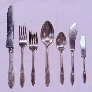 May include: A set of vintage silverware, including a knife, two forks, two spoons, and two butter knives. The silverware is silver-colored with ornate handle detailing. The arrangement is on a light purple background, showcasing the complete dining set.