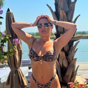 May include: A leopard print bikini with a gold clasp. The two-piece swimsuit features a high-cut bottom and a triangle top. The person is wearing sunglasses and a watch. The background includes a palm tree, flowers, and a body of water.