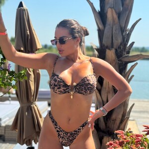 May include: A two-piece leopard print bikini with a gold clasp. The bikini top has thin straps and a sweetheart neckline. The bottoms are high-cut. The person is wearing sunglasses and a bracelet. The background includes a beach umbrella and palm trees.