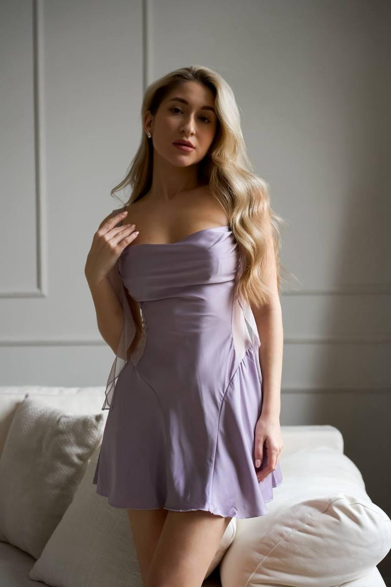 Puede incluir: Un minivestido lila sin tirantes con una falda fluida. El vestido presenta un corpi&ntilde;o drapeado y detalles de tela transparente. La modelo tiene el pelo largo y rubio y est&aacute; frente a un sof&aacute; blanco.
