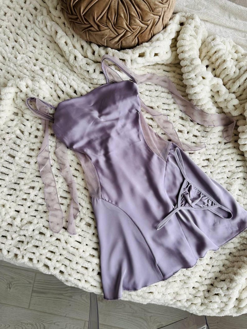 Puede incluir: Un conjunto de lencer&iacute;a de sat&eacute;n lavanda, que incluye un camis&oacute;n y un tanga a juego. El camis&oacute;n presenta delicadas correas y paneles de malla transparente. El conjunto se exhibe sobre una manta de punto grueso blanca.