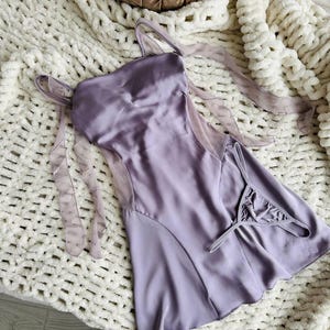 Puede incluir: Un conjunto de lencer&iacute;a de sat&eacute;n lavanda, que incluye un camis&oacute;n y un tanga a juego. El camis&oacute;n presenta tirantes finos y paneles de malla transparente. El conjunto se exhibe sobre una manta blanca de punto grueso.