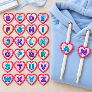 May include: A light blue hooded sweatshirt with two white drawstring cords, each featuring a pink heart-shaped charm with a letter. Beside the sweatshirt are twenty-six individual heart-shaped charms, each displaying a unique letter from A to Z.