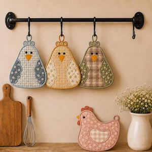 May include: Three quilted chicken-shaped pot holders in blue, yellow, and green gingham and floral patterns hang from a black metal rack. A fourth pink floral chicken-shaped pot holder sits on a surface next to a wooden board and a wire whisk.