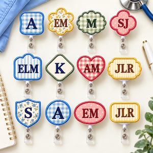 May include: Twelve retractable badge reels in various shapes and patterns, including gingham, floral, and polka dot designs. Each reel features embroidered letters or initials such as A, EM, M, SJ, ELM, K, AM, and JLR in dark thread.