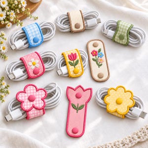May include: A collection of cable organizers featuring various floral and gingham patterns. These fabric straps use snap buttons to secure coiled white charging cables. Designs include flower-shaped clips, embroidered tulips, and colorful checkered prints.