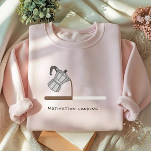 May include: A pale pink sweatshirt with a crew neck and long sleeves. The front displays a graphic of a coffee maker pouring coffee into a loading bar, with the text "MOTIVATION LOADING..".