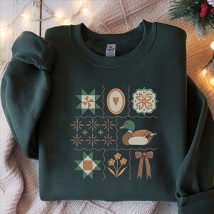 May include: Dark green sweatshirt featuring a patchwork design with various geometric shapes, a heart, and a duck illustration. The design includes stars, flowers, and a bow. The sweatshirt is made of a soft material.