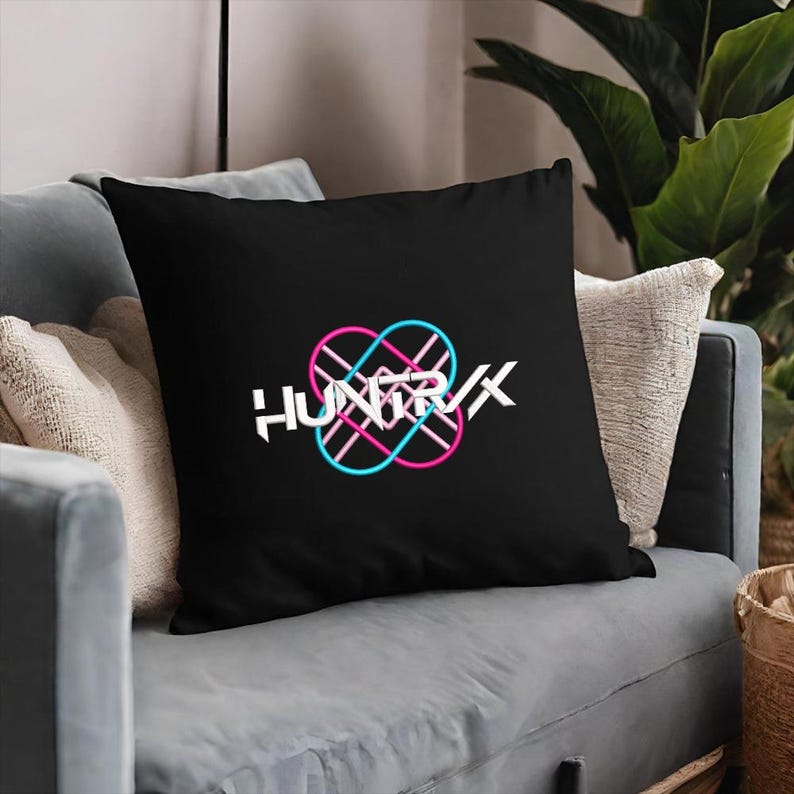 Puede incluir: Coj&iacute;n cuadrado negro con la palabra "HUNTRX" en blanco, con un dise&ntilde;o entrelazado rosa y azul. El coj&iacute;n est&aacute; sobre un sof&aacute; gris con otros cojines y una planta en el fondo.
