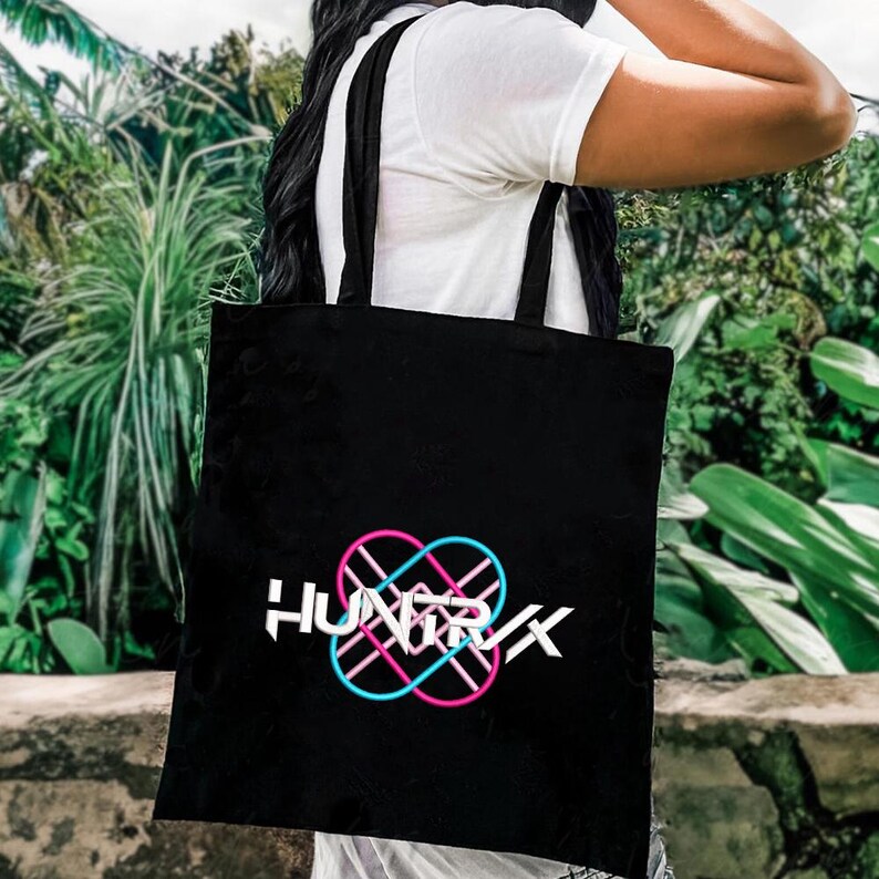 Puede incluir: Bolso tote negro con la palabra "HUNTRX" en blanco, con un dise&ntilde;o de s&iacute;mbolo de infinito rosa y azul. El bolso tiene correas negras y se lleva sobre el hombro de una persona. El fondo es follaje verde borroso.