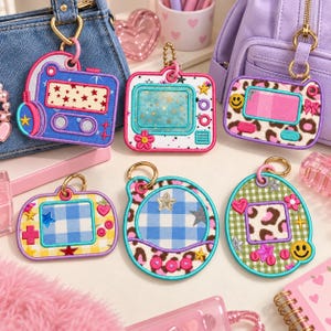 May include: A collection of six embroidered keychains featuring various shapes and patterns, including a blue cassette player, a white television, a leopard print device, and three rounded digital game consoles with gingham and leopard accents.